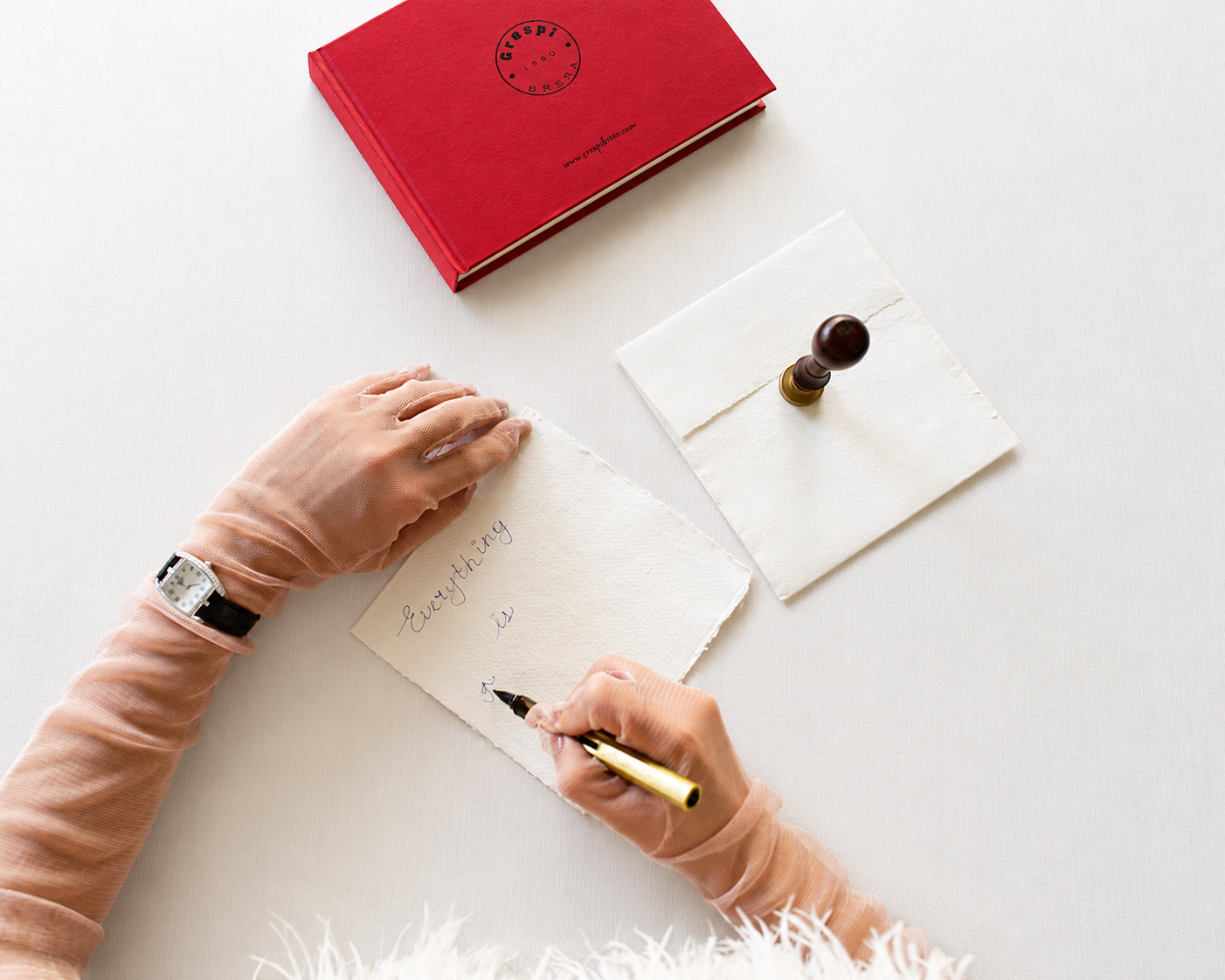 Hands writing Everything is on handmade paper beside a red book and wax seal